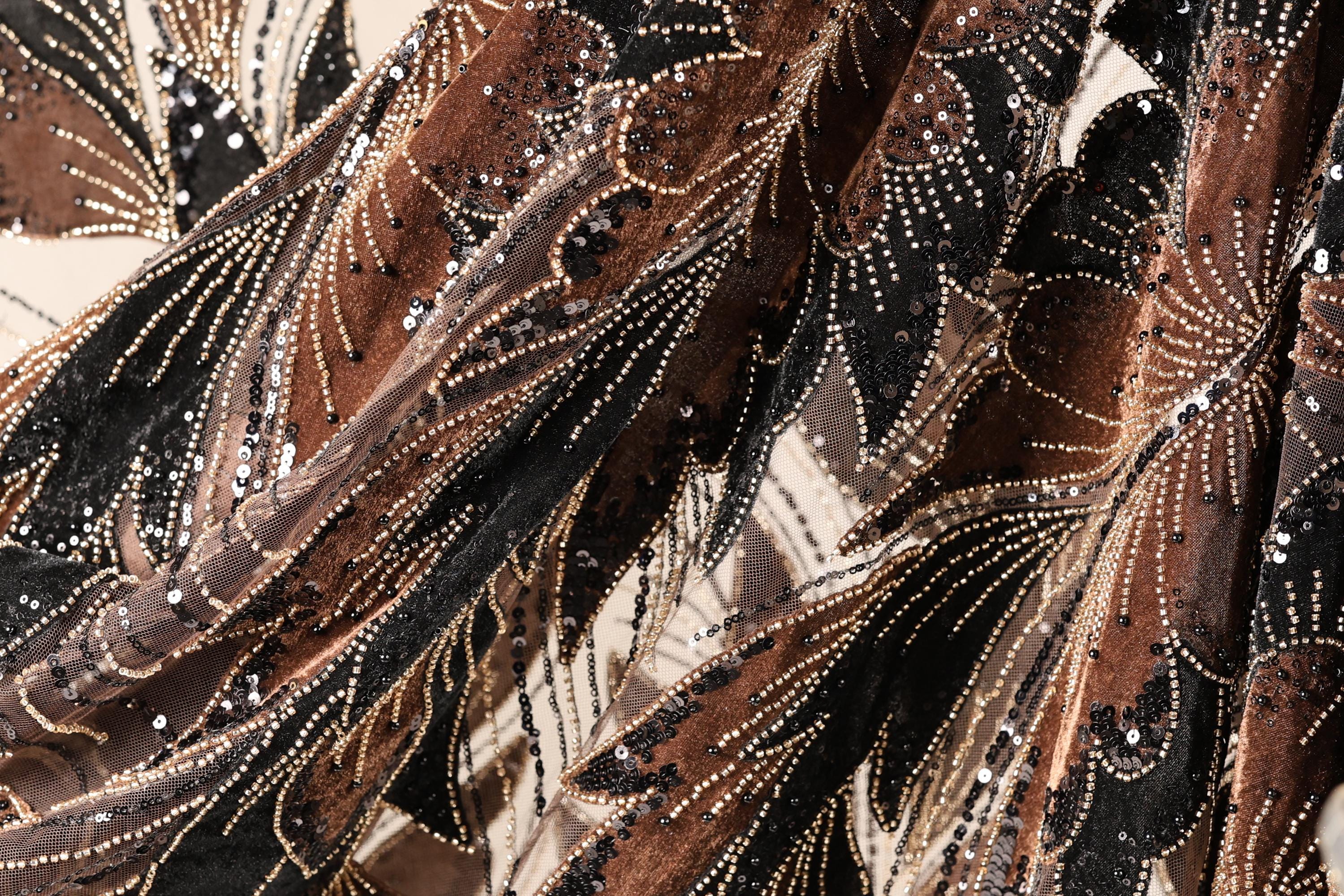 The image is a close-up of a fabric with a floral pattern, featuring a mix of brown, black, and gold colors. The fabric appears to be made of a shiny material, possibly sequin or lace, and has a textured, flowing appearance.