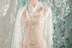 A mannequin wearing a white lace dress with intricate floral embroidery, set against a backdrop of shimmering silver and green sequins.