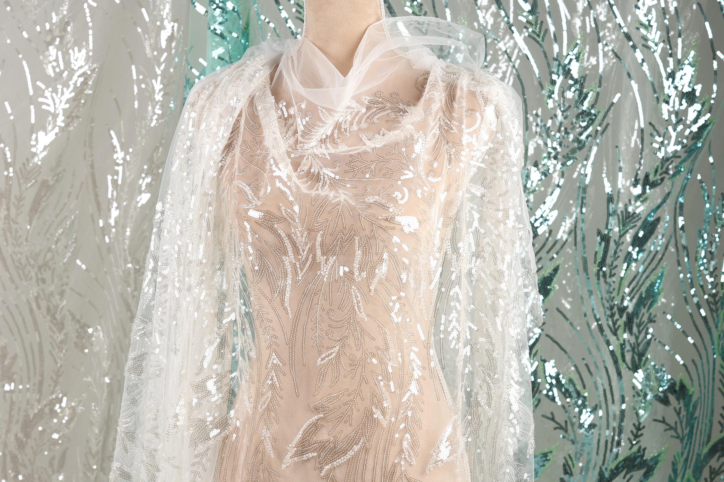 A mannequin wearing a white lace dress with intricate floral embroidery, set against a backdrop of shimmering silver and green sequins.