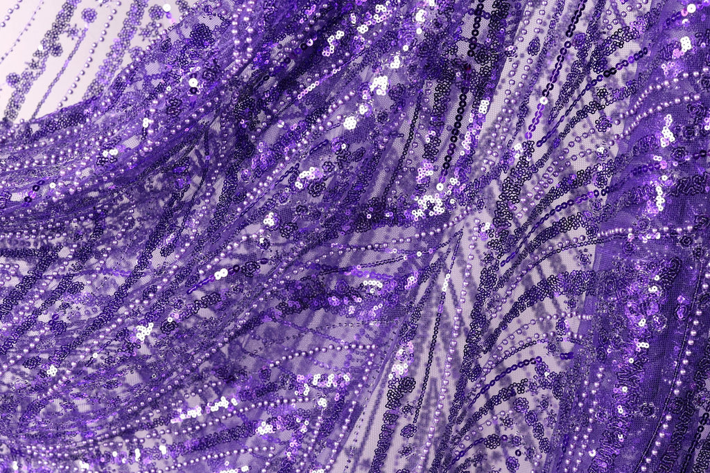 The image is a close-up view of a fabric with a purple and white pattern, featuring a lot of sequins and beads. The fabric appears to be made of a shiny, glittery material, giving it a festive and celebratory appearance.