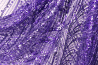 The image is a close-up view of a fabric with a purple and white pattern, featuring a lot of sequins and beads. The fabric appears to be made of a shiny, glittery material, giving it a festive and celebratory appearance.