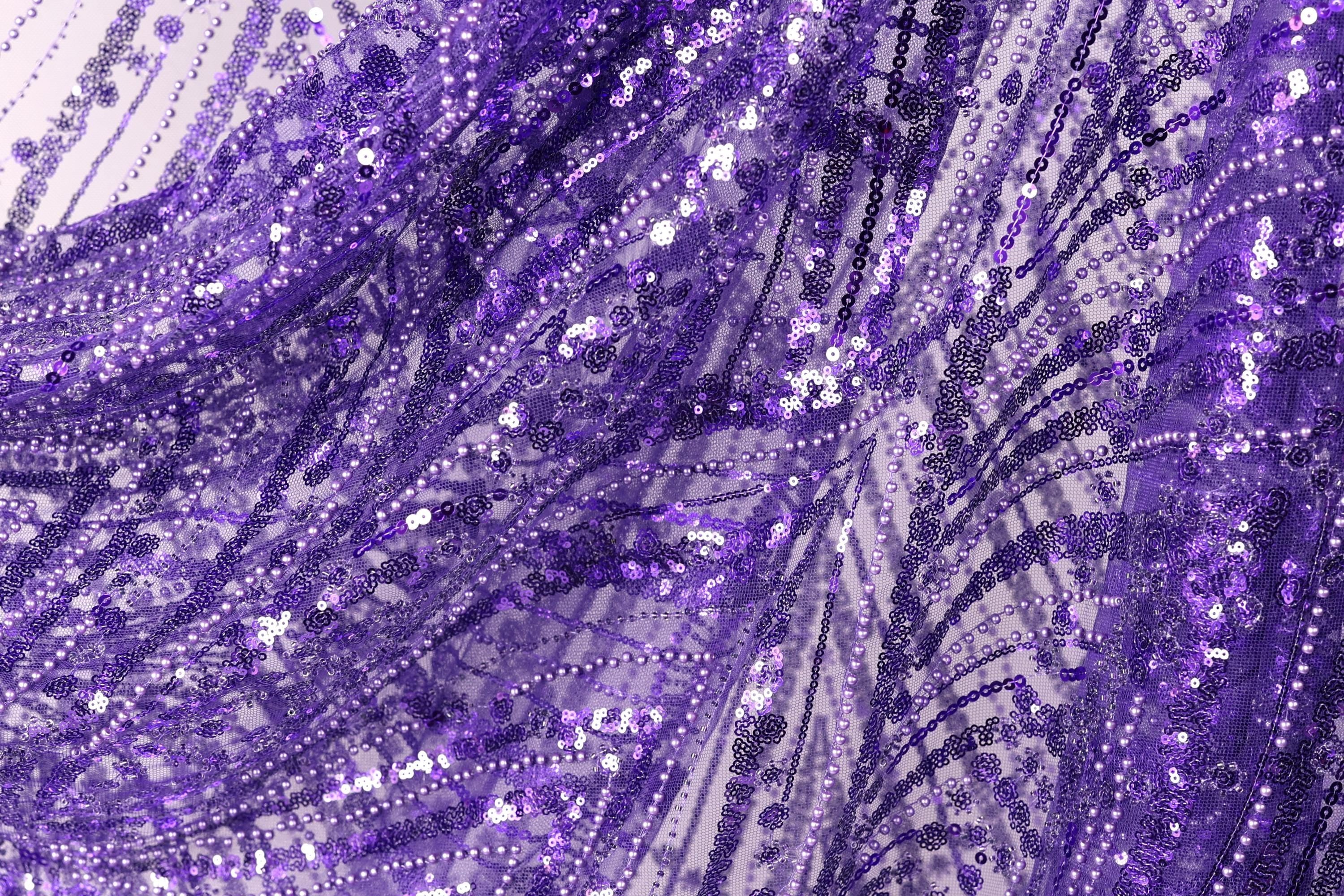 The image is a close-up view of a fabric with a purple and white pattern, featuring a lot of sequins and beads. The fabric appears to be made of a shiny, glittery material, giving it a festive and celebratory appearance.