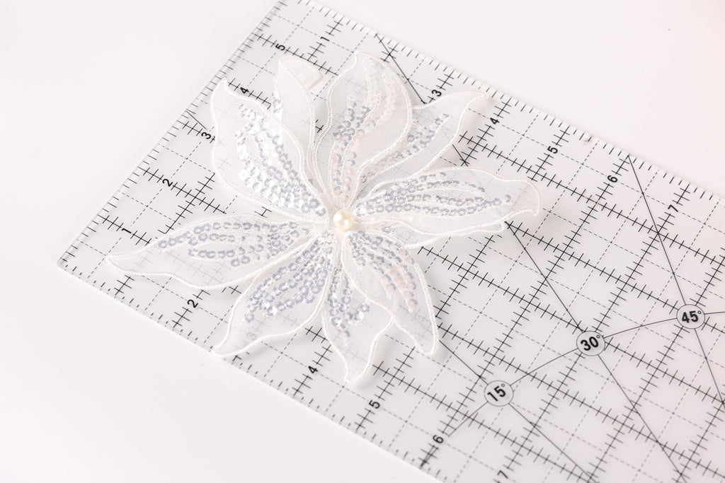 A white sequin flower is placed on a clear ruler with measurement markings.