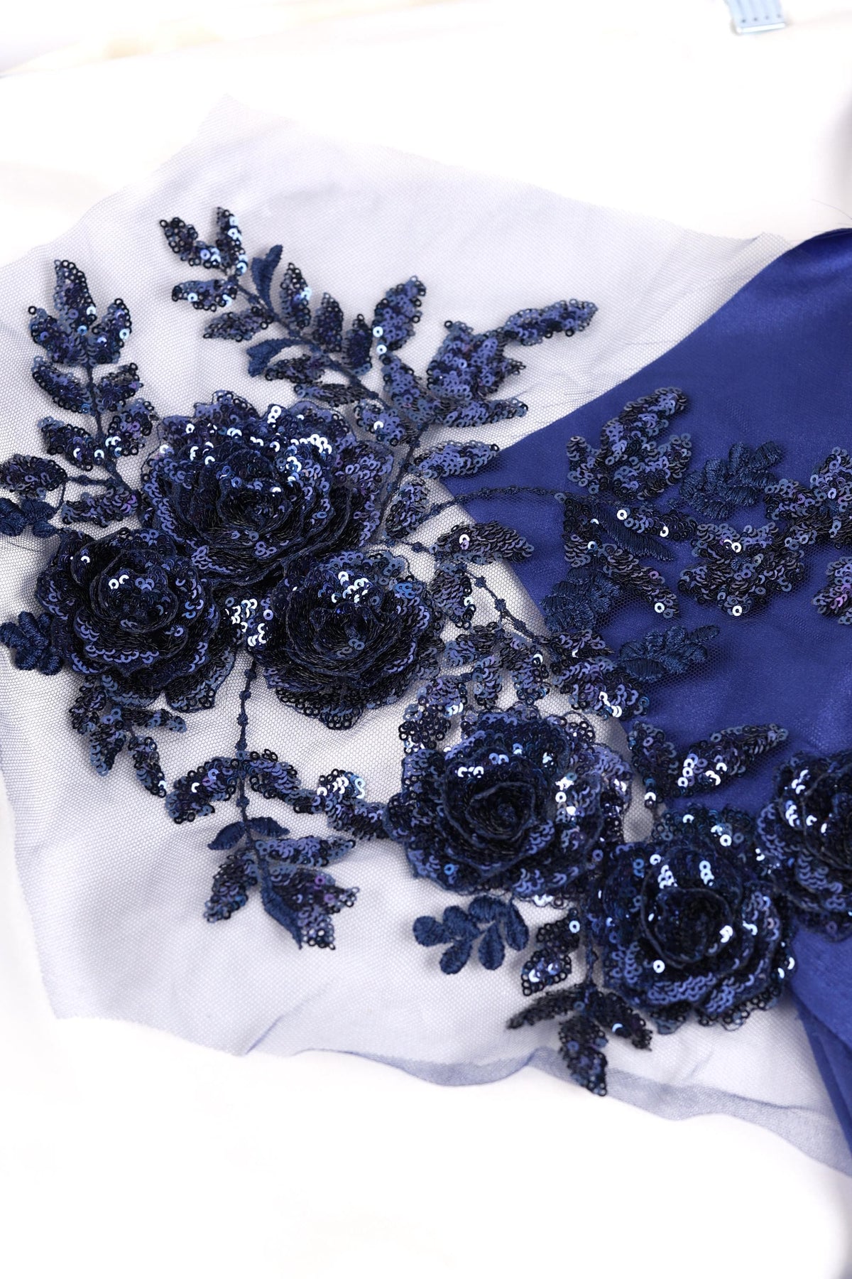 a close-up view of a fabric with a floral pattern, including blue sequin flowers and leaves. The fabric appears to be a light blue color, and the intricate details of the floral design are clearly visible.