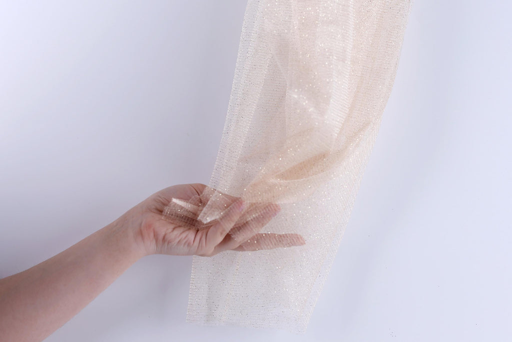 A hand is holding a piece of beige, shimmering fabric against a white background.