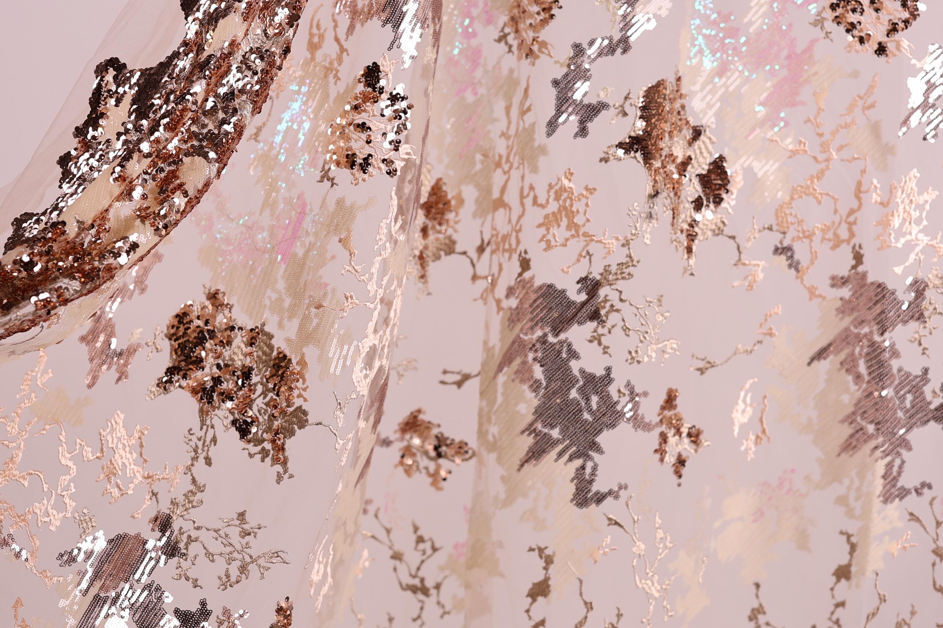 The image is a close-up of a fabric with a metallic, sequined pattern in shades of pink, brown, and gold. The fabric appears to be a sheer or sheer-like material, with a textured and shiny appearance.