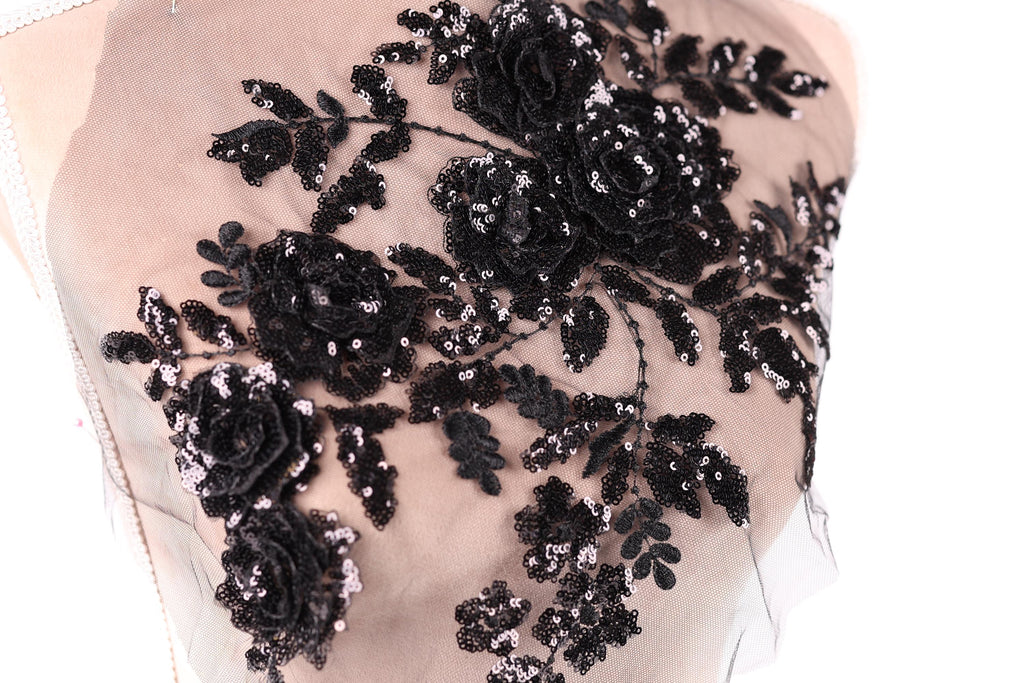 a close-up view of a delicate, intricate black lace design with a floral pattern. The lace appears to be made of sheer fabric, giving it a delicate and elegant appearance. The design features a variety of black flowers and leaves, which are intricately sewn onto the fabric, creating a visually striking and detailed piece of textile art.