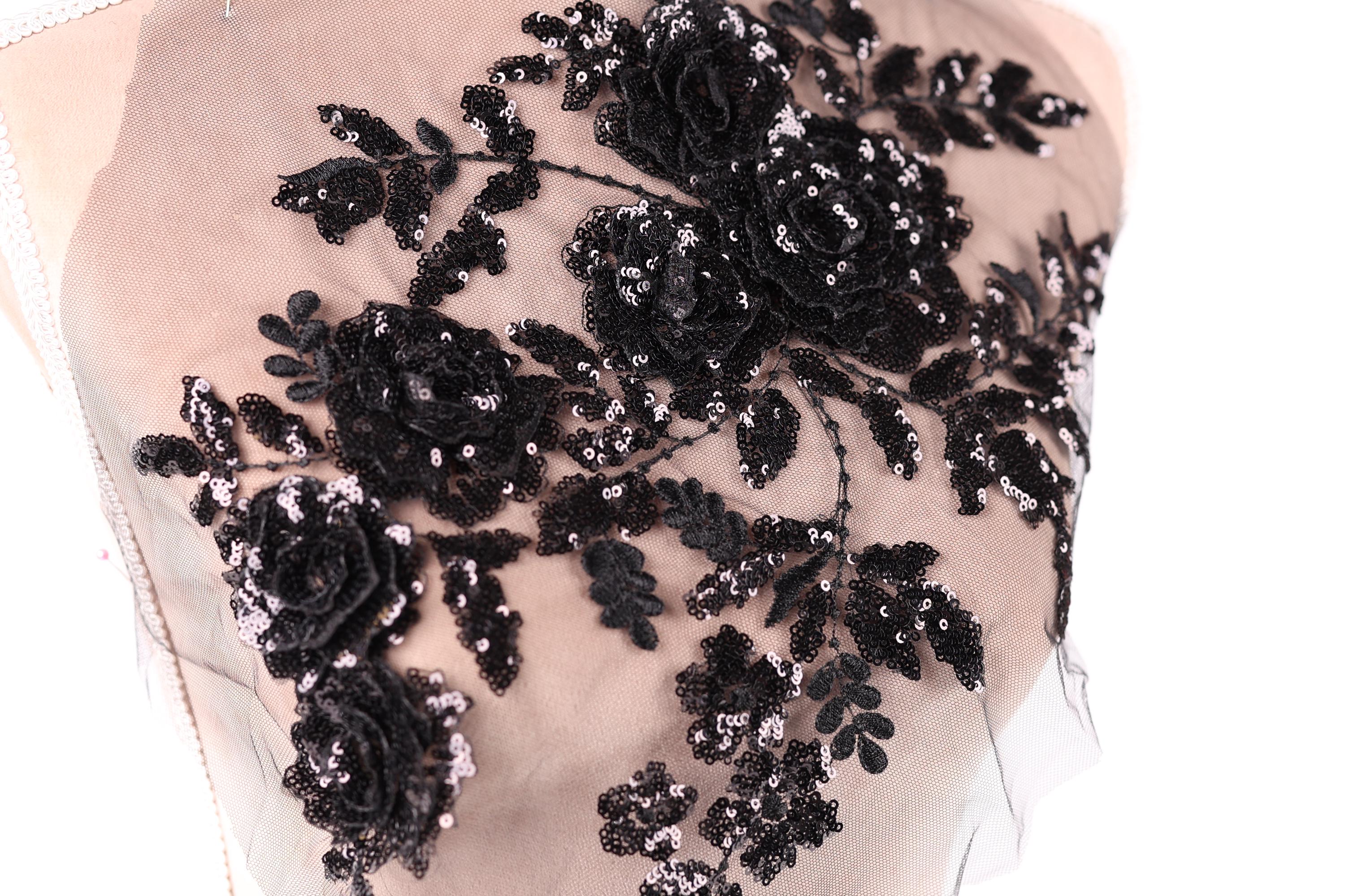 a close-up view of a delicate, intricate black lace design with a floral pattern. The lace appears to be made of sheer fabric, giving it a delicate and elegant appearance. The design features a variety of black flowers and leaves, which are intricately sewn onto the fabric, creating a visually striking and detailed piece of textile art.