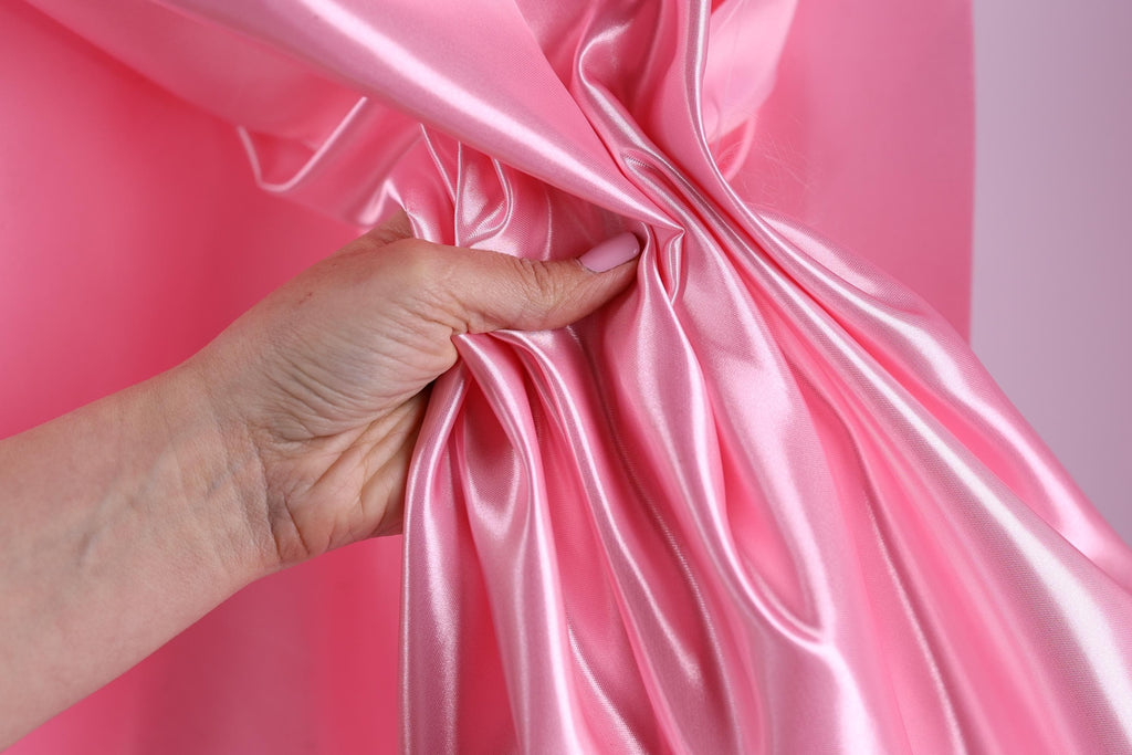 A hand is holding a pink satin fabric, with the fabric being crumpled and wrinkled.
