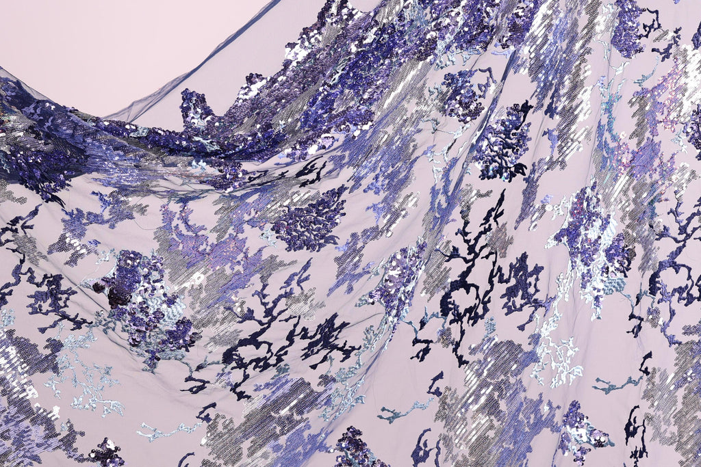 The image is a close-up view of a fabric with a floral pattern in shades of purple and blue. The fabric appears to be made of sequins or other decorative materials, creating a visually striking and intricate design.