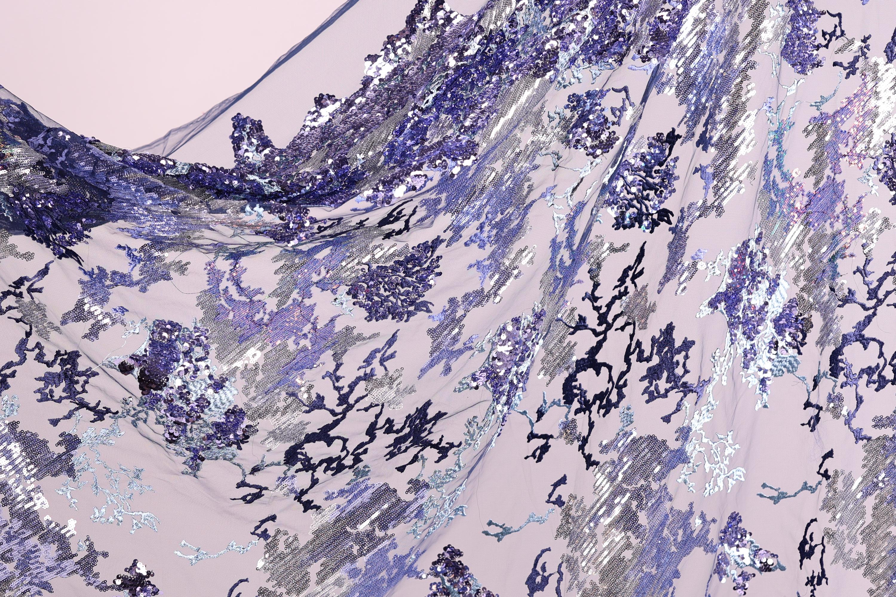 The image is a close-up view of a fabric with a floral pattern in shades of purple and blue. The fabric appears to be made of sequins or other decorative materials, creating a visually striking and intricate design.