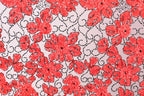 a close-up view of a fabric with a repeating pattern of red flowers against a light background. The flowers are intricately designed and appear to be made of a shiny material, giving them a sparkling appearance. The pattern is composed of numerous small red flowers arranged in a visually striking manner, creating a vibrant and eye-catching design.