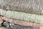 A close-up image of a pile of fabric with sequins and lace-like patterns, showcasing various shades of gray, pink, and green.