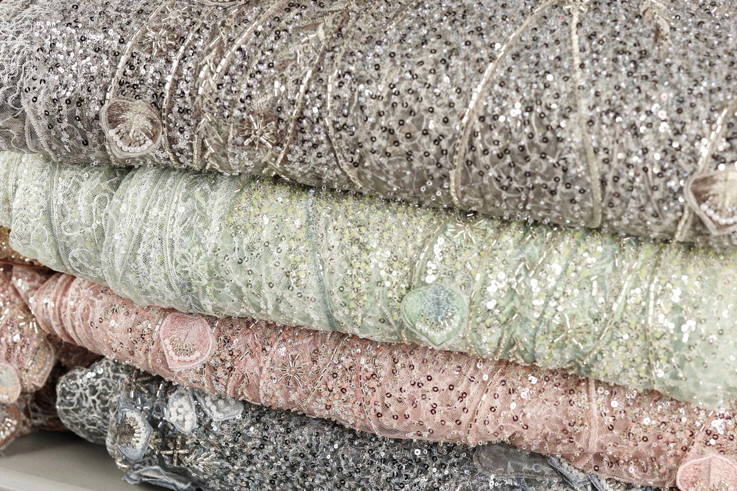 A close-up image of a pile of fabric with sequins and lace-like patterns, showcasing various shades of gray, pink, and green.