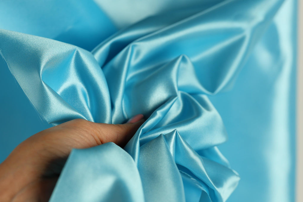 A hand is holding a piece of blue satin fabric, which appears to be a close-up view of the material.
