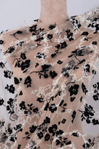a mannequin wearing a sheer fabric with a black floral pattern. The fabric appears to be a sheer lace or sheer chiffon, with a delicate and intricate design of black flowers and leaves. The mannequin is positioned in a way that highlights the fabric's texture and pattern, creating a visually appealing display.