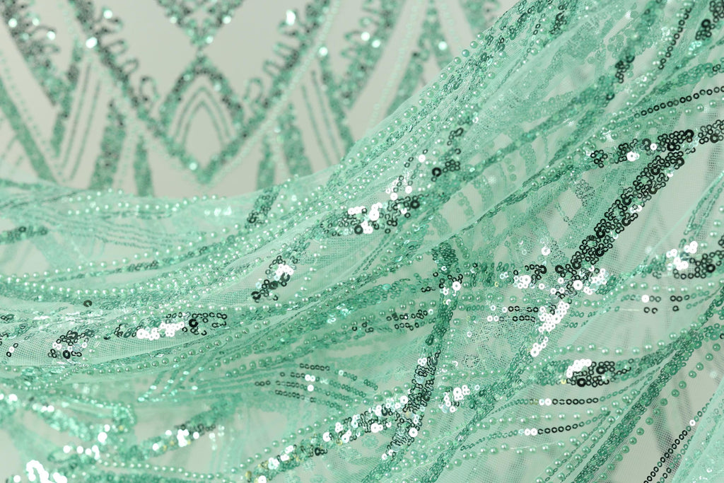 The image is a close-up of a fabric with a green and silver sequin pattern, featuring a mix of small and large sequins arranged in a swirling, wavy design.