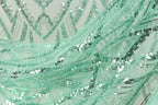 The image is a close-up of a fabric with a green and silver sequin pattern, featuring a mix of small and large sequins arranged in a swirling, wavy design.