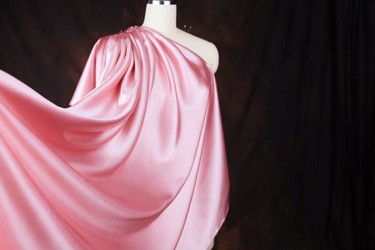 A mannequin wearing a flowing, one-shoulder pink satin dress against a dark background.