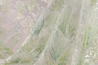 a close-up view of a delicate, intricately embroidered fabric with a floral pattern. The fabric appears to be made of a light-colored material, possibly silk or chiffon, and is adorned with small, colorful beads or sequins that add a touch of sparkle and detail to the design. The fabric is folded and crumpled, giving it a soft, flowing appearance.