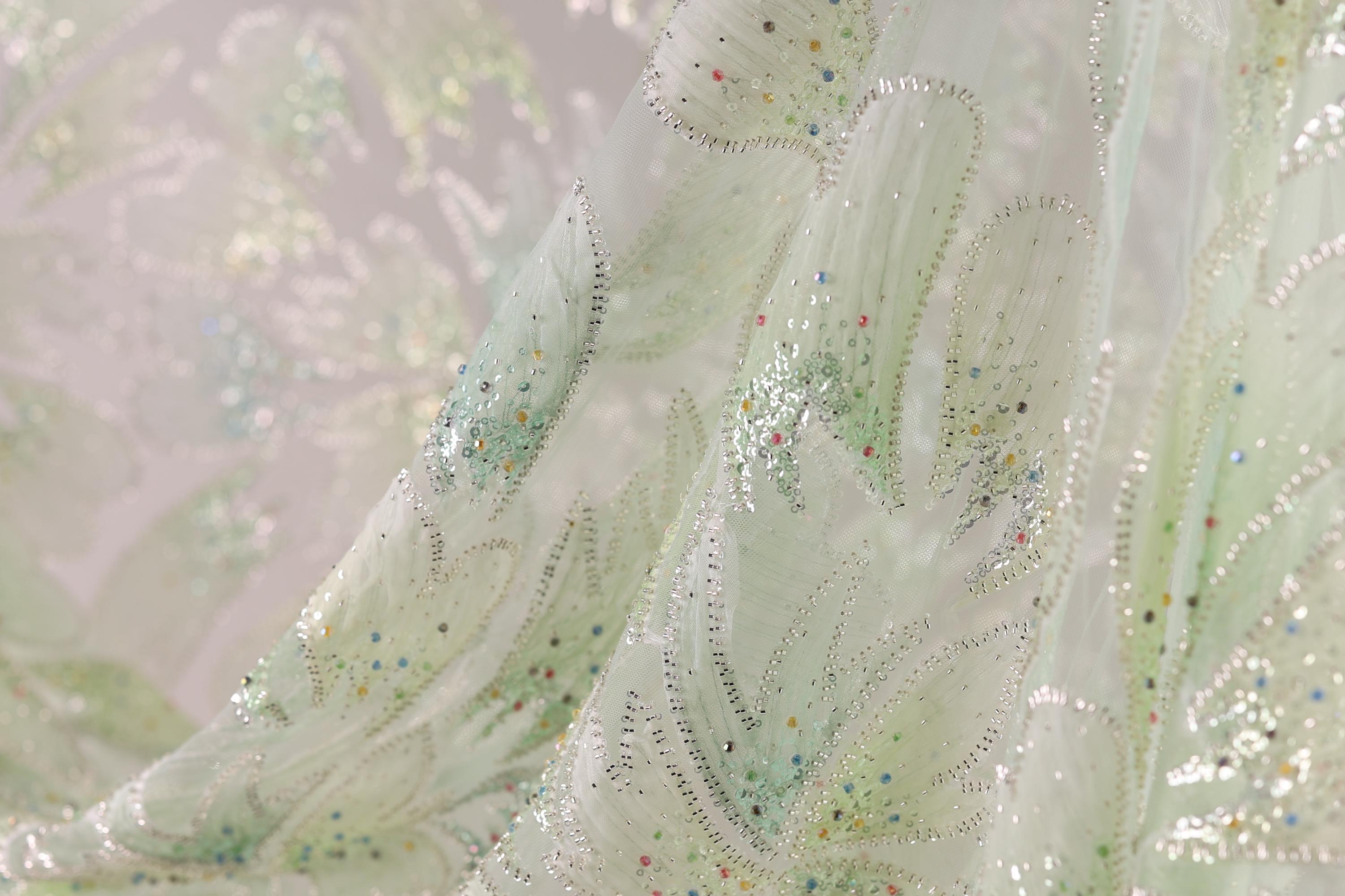 a close-up view of a delicate, intricately embroidered fabric with a floral pattern. The fabric appears to be made of a light-colored material, possibly silk or chiffon, and is adorned with small, colorful beads or sequins that add a touch of sparkle and detail to the design. The fabric is folded and crumpled, giving it a soft, flowing appearance.