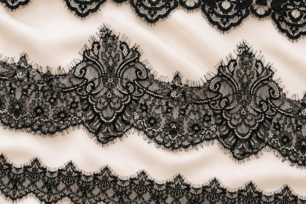 a close-up view of a delicate, intricate black lace pattern on a soft, light-colored fabric. The lace appears to be made of silk and is adorned with ornate, floral-inspired designs. The fabric has a smooth, flowing texture, and the lace is arranged in a repeating pattern across the entire surface.