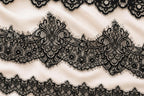 a close-up view of a delicate, intricate black lace pattern on a soft, light-colored fabric. The lace appears to be made of silk and is adorned with ornate, floral-inspired designs. The fabric has a smooth, flowing texture, and the lace is arranged in a repeating pattern across the entire surface.