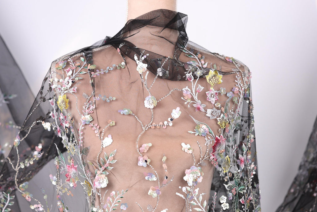 a close-up view of a sheer, black fabric with a floral pattern. The fabric appears to be made of sheer material, giving it a delicate and airy appearance. The floral pattern consists of various flowers and leaves, creating a visually appealing and intricate design. The fabric is displayed on a mannequin, which is positioned in the center of the image. The background of the image is white, which contrasts with the black fabric and allows the details of the floral pattern to stand out.