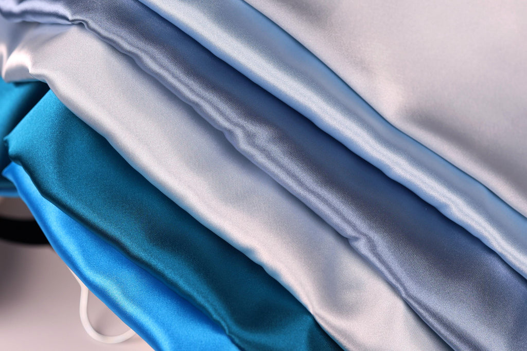 A close-up image of various colored satin fabric, including shades of blue, gray, and white, arranged in a layered fashion.