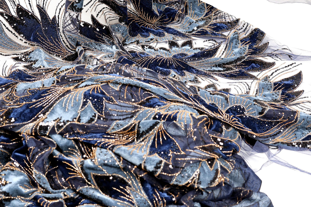 a close-up view of a fabric with a blue and gold floral pattern. The fabric appears to be made of a shiny material, possibly sequin or lace, and has a textured, crumpled appearance.