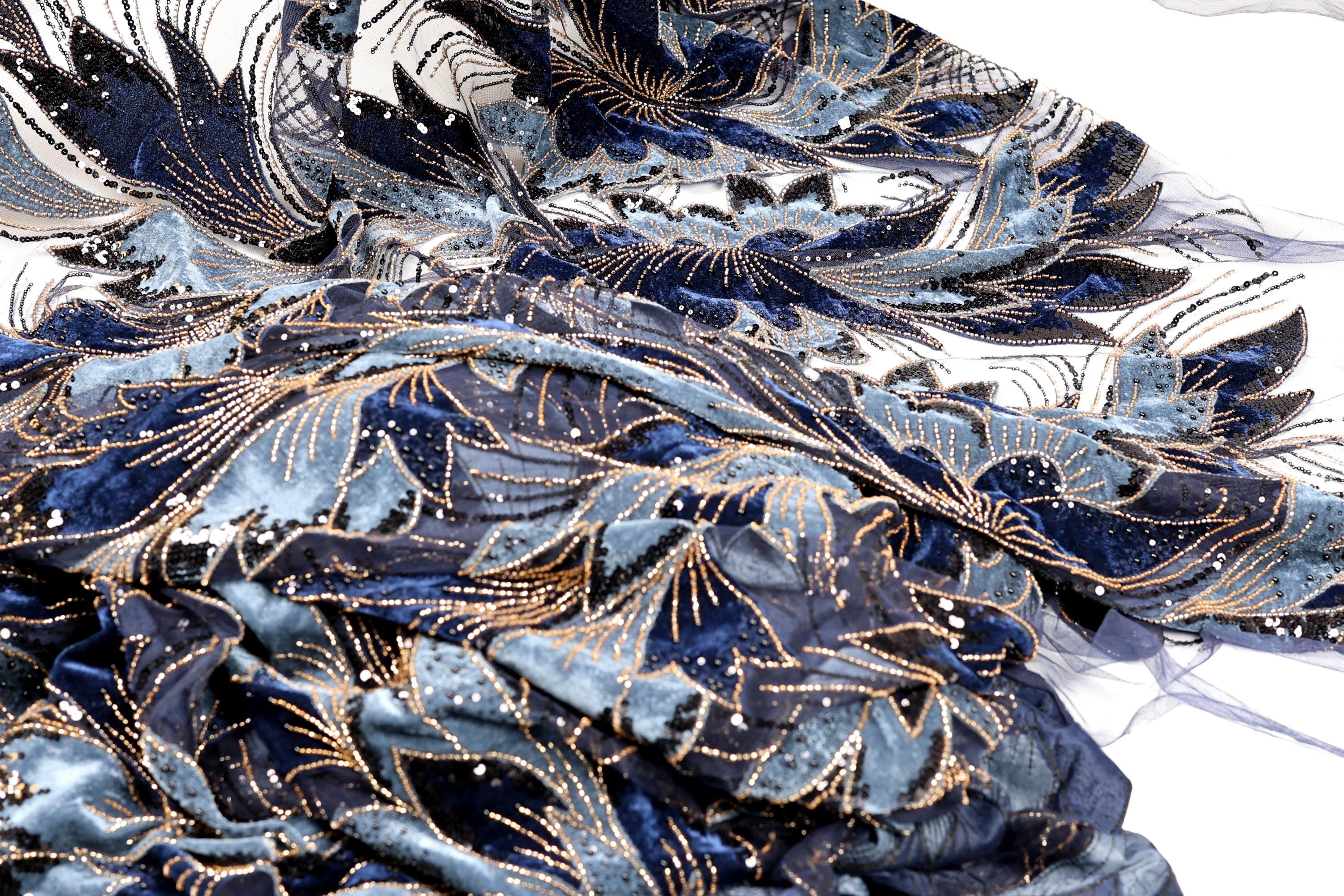 a close-up view of a fabric with a blue and gold floral pattern. The fabric appears to be made of a shiny material, possibly sequin or lace, and has a textured, crumpled appearance.