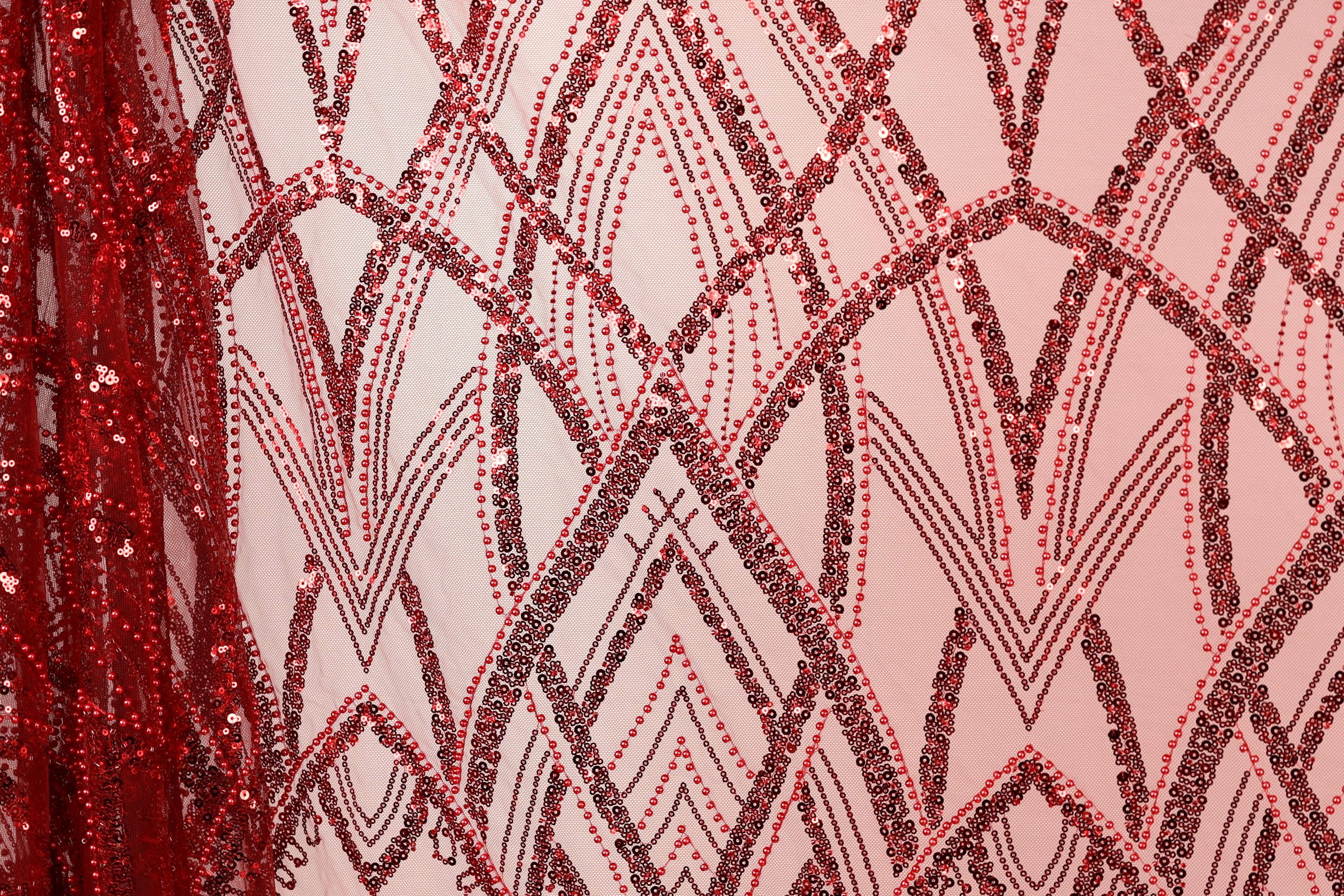 a close-up view of a fabric with a repeating pattern of red and black sequin designs. The pattern consists of various shapes and lines, creating a visually striking and intricate design. The fabric appears to be a light pink color, which contrasts with the red sequins and adds to the overall visual appeal of the image.
