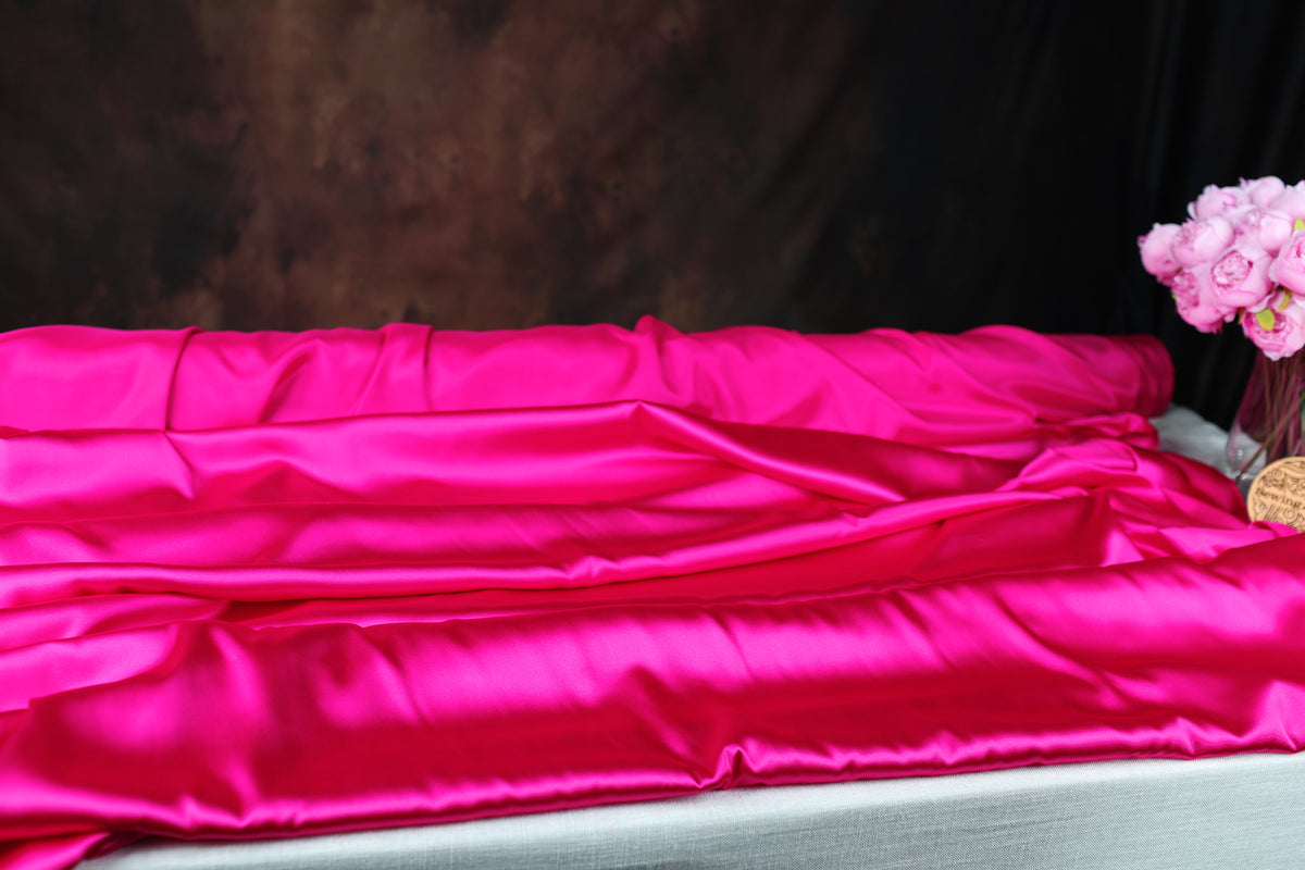 a large, vibrant pink satin fabric draped over a white surface, with a vase of pink flowers placed next to it.