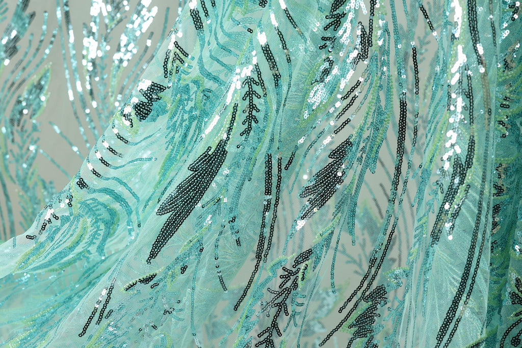 a close-up view of a fabric with a pattern of sequins and beads in shades of blue and green. The fabric appears to be made of sheer material, giving it a delicate and flowing appearance. The pattern on the fabric is intricate and detailed, with various lines and shapes creating a visually striking design.