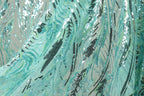 a close-up view of a fabric with a pattern of sequins and beads in shades of blue and green. The fabric appears to be made of sheer material, giving it a delicate and flowing appearance. The pattern on the fabric is intricate and detailed, with various lines and shapes creating a visually striking design.