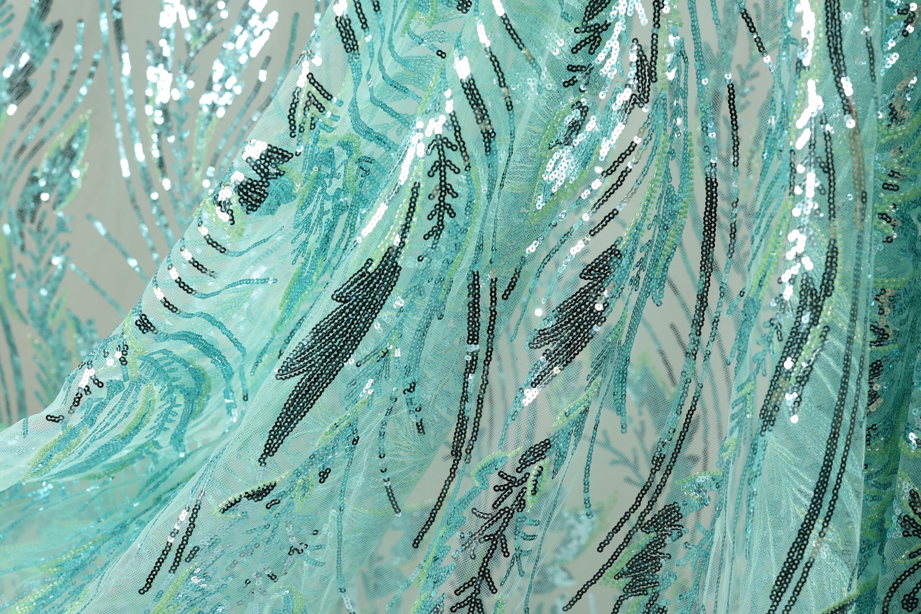 a close-up view of a fabric with a pattern of sequins and beads in shades of blue and green. The fabric appears to be made of sheer material, giving it a delicate and flowing appearance. The pattern on the fabric is intricate and detailed, with various lines and shapes creating a visually striking design.
