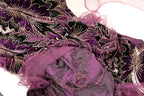 The image is a close-up of a fabric with a floral pattern in shades of purple and black, featuring gold and white embellishments.