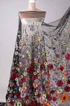 a mannequin wearing a dress with a sheer, sheer-like fabric that is decorated with a variety of colorful flowers. The dress has a long, flowing skirt that extends outwards, creating a sense of movement and elegance. The flowers are arranged in a way that covers the entire dress, adding a vibrant and eye-catching element to the overall design.