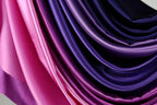 a close-up view of a fabric with a gradient of colors, transitioning from pink to purple. The fabric appears to be made of satin or a similar material, with a smooth and shiny texture. The colors are vibrant and distinct, creating a visually striking and eye-catching display.
