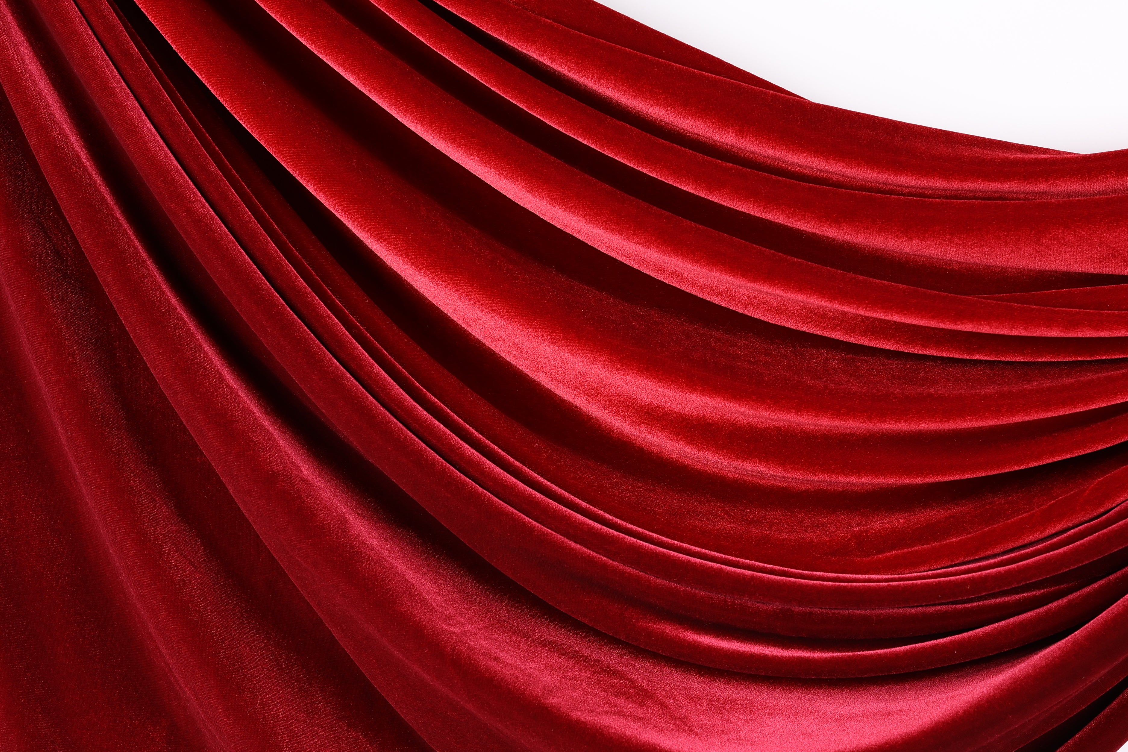 a close-up view of a red velvet fabric, with its smooth and shiny texture visible. The fabric appears to be draped or flowing, creating a sense of depth and movement. The color of the fabric is rich and vibrant, with a deep red hue that contrasts with the white background. The fabric's texture is smooth and shiny, adding to its visual appeal.
