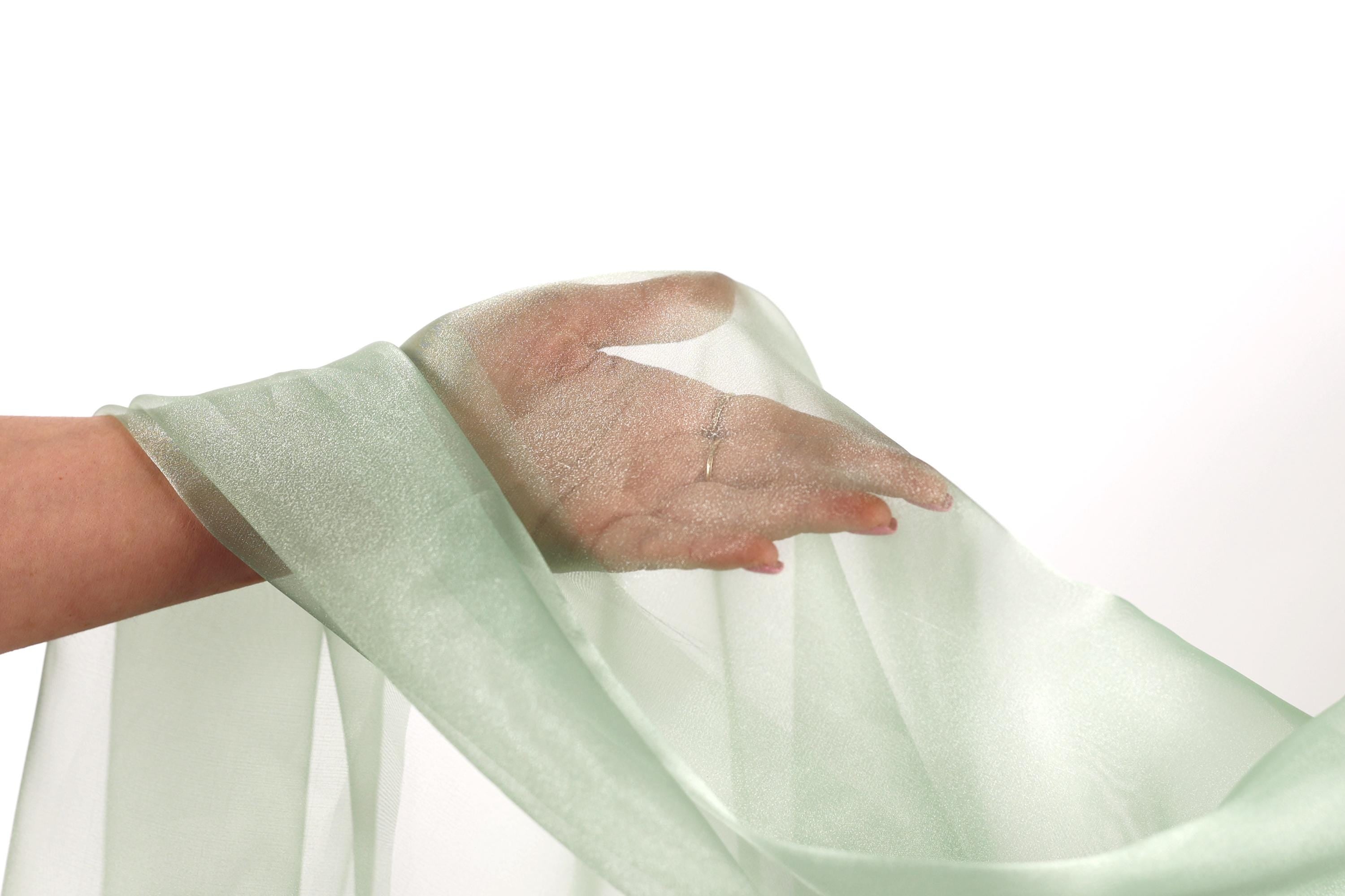 A hand is holding a green fabric, possibly a sheer or sheer-like material, against a white background.