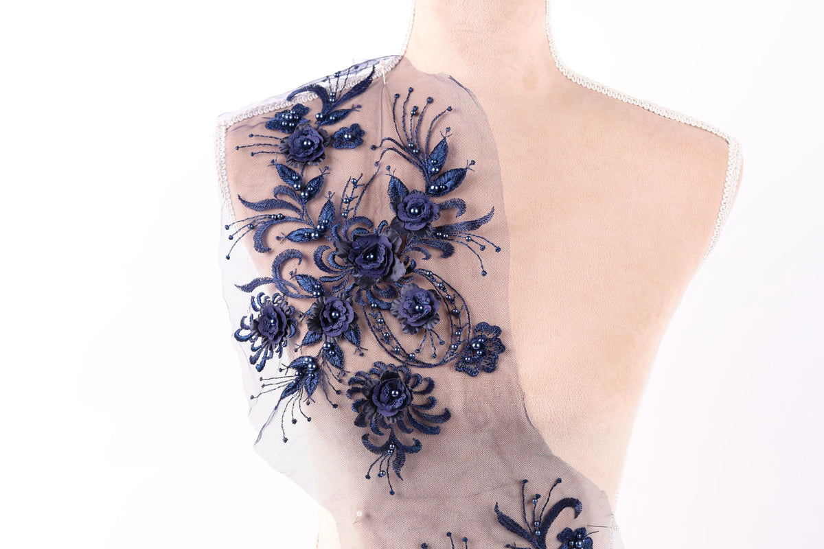 a mannequin with a decorative fabric piece on its shoulder. The fabric piece is adorned with a floral pattern in shades of blue, including various types of flowers and leaves. The mannequin is positioned against a plain white background, allowing the intricate details of the fabric piece to be the focal point of the image.