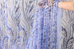 a hand holding a piece of fabric with a floral pattern in shades of blue and purple. The fabric appears to be made of sequin material and has a delicate, intricate design.