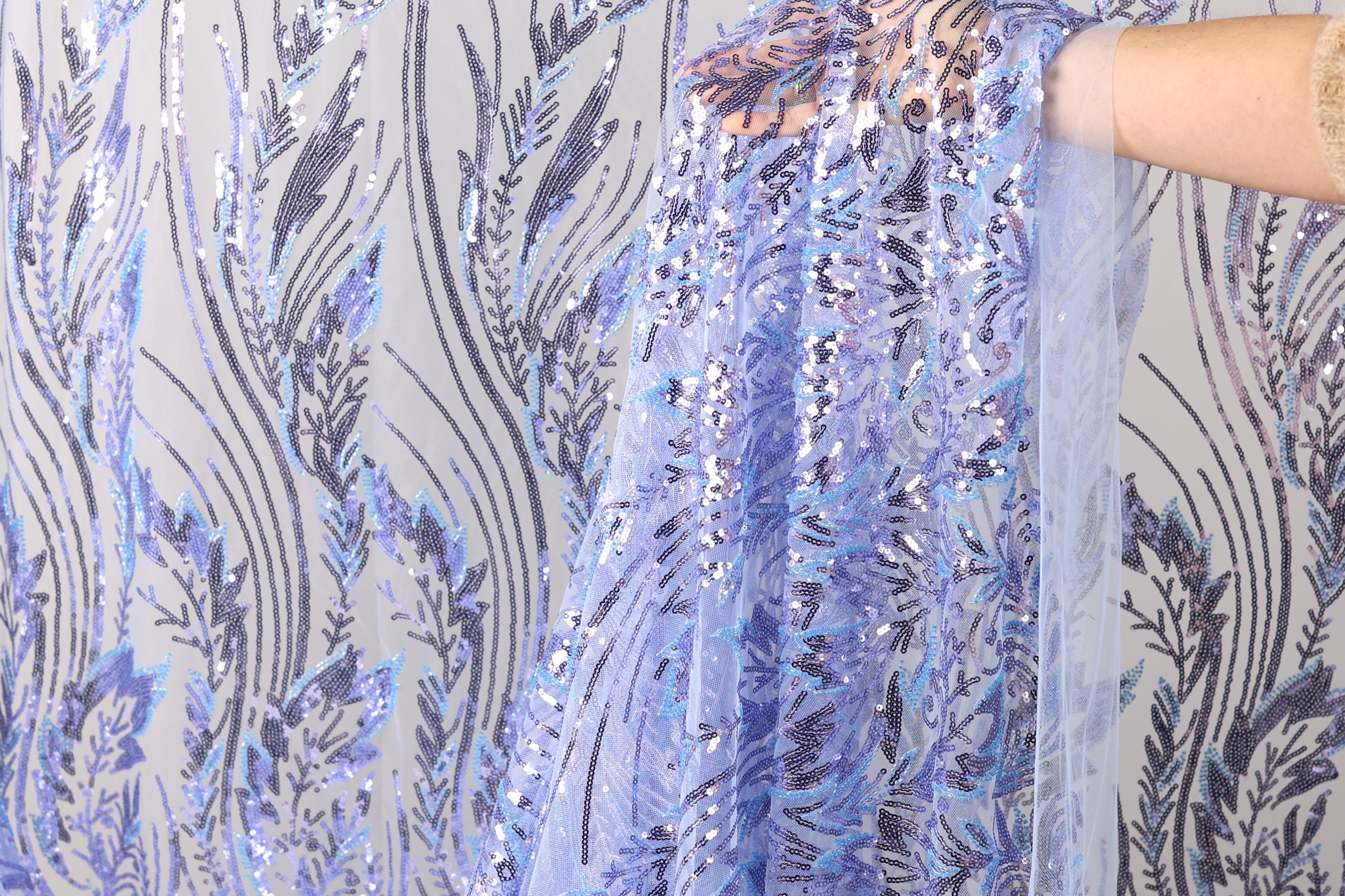 a hand holding a piece of fabric with a floral pattern in shades of blue and purple. The fabric appears to be made of sequin material and has a delicate, intricate design.