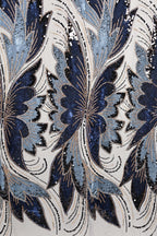 a close-up view of a fabric with a repeating pattern of blue and white butterfly-like shapes, adorned with sequins and beads. The pattern is intricate and detailed, showcasing the craftsmanship and artistry involved in creating the fabric.