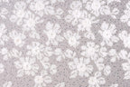 a close-up view of a fabric with a floral pattern. The fabric appears to be made of a light-colored material, possibly lace or a similar type of material. The pattern consists of numerous small white flowers or petals scattered across the fabric, creating a visually appealing and intricate design.