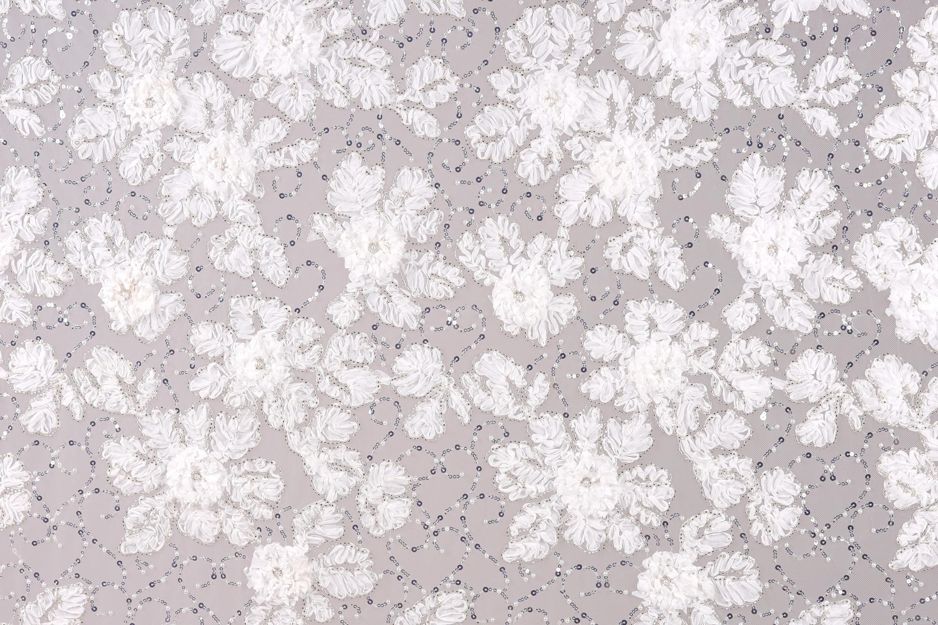 a close-up view of a fabric with a floral pattern. The fabric appears to be made of a light-colored material, possibly lace or a similar type of material. The pattern consists of numerous small white flowers or petals scattered across the fabric, creating a visually appealing and intricate design.