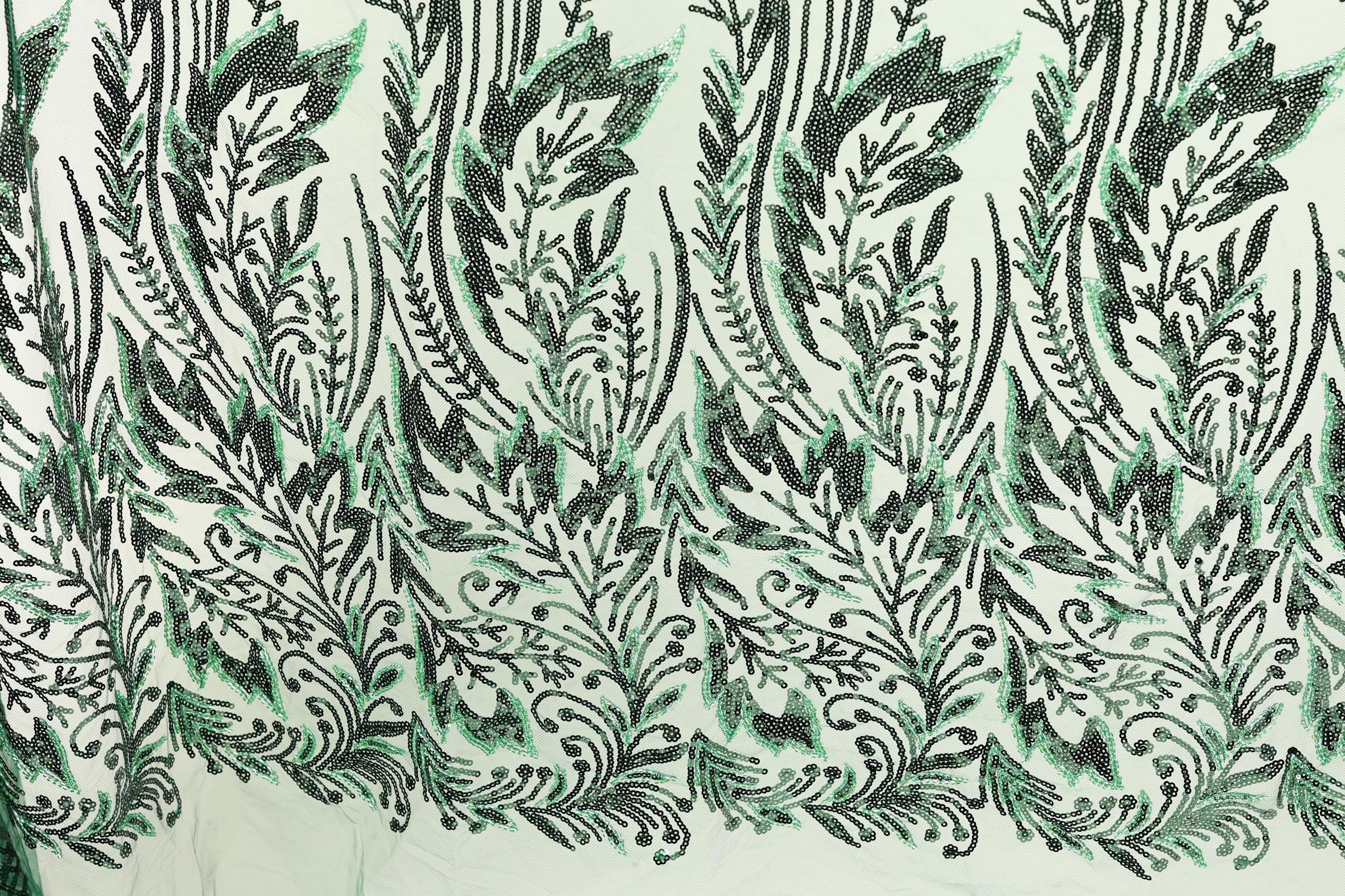 a close-up view of a fabric with a repeating pattern of green and black leaf-like designs. The pattern is intricate and detailed, with the leaves appearing to be made of sequins or other embellishments. The fabric has a textured appearance, suggesting it could be a type of clothing or textile.