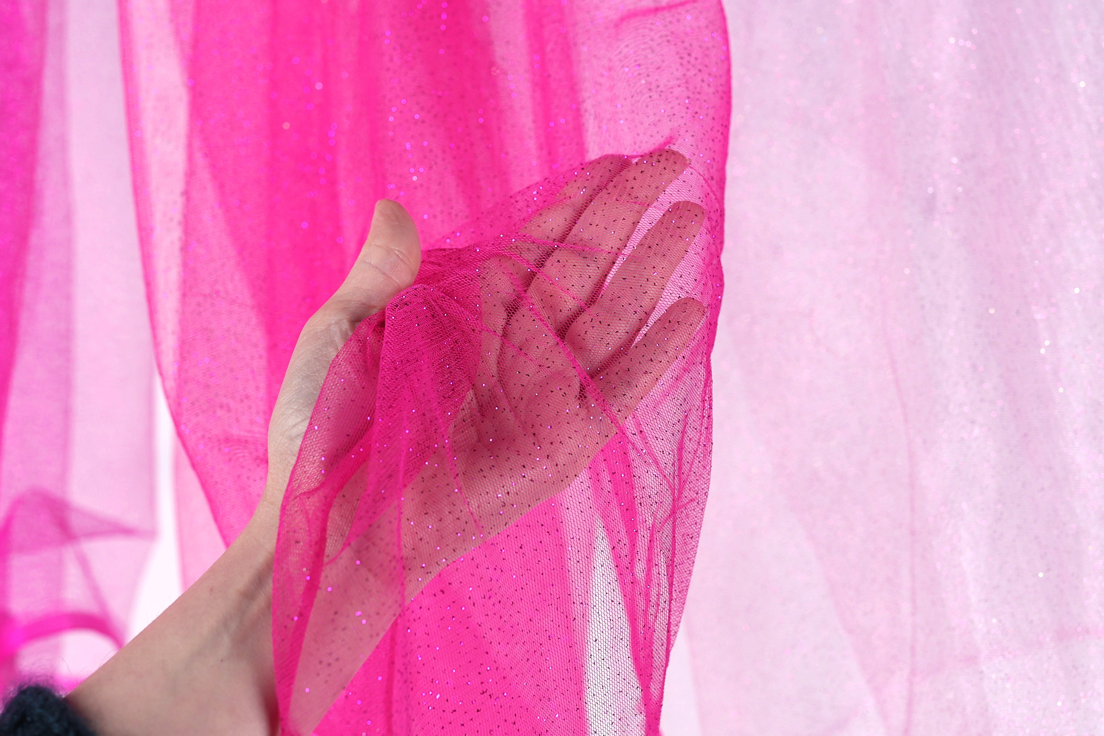 A hand is holding a pink, sparkly fabric against a pink background.