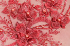 The image is a close-up of a delicate, intricate piece of red embroidery featuring a floral pattern with red flowers and small pearls.